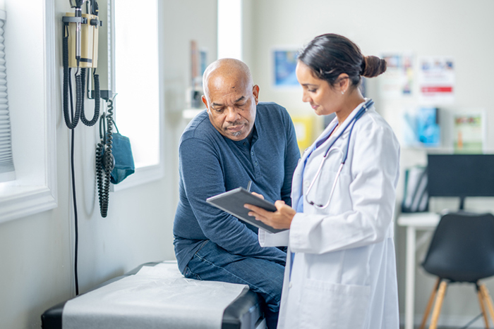 A senior gentleman of African decent sits up on an exam table during a routine check-up. His female doctor is holding out a tablet as they review some recent test results together.